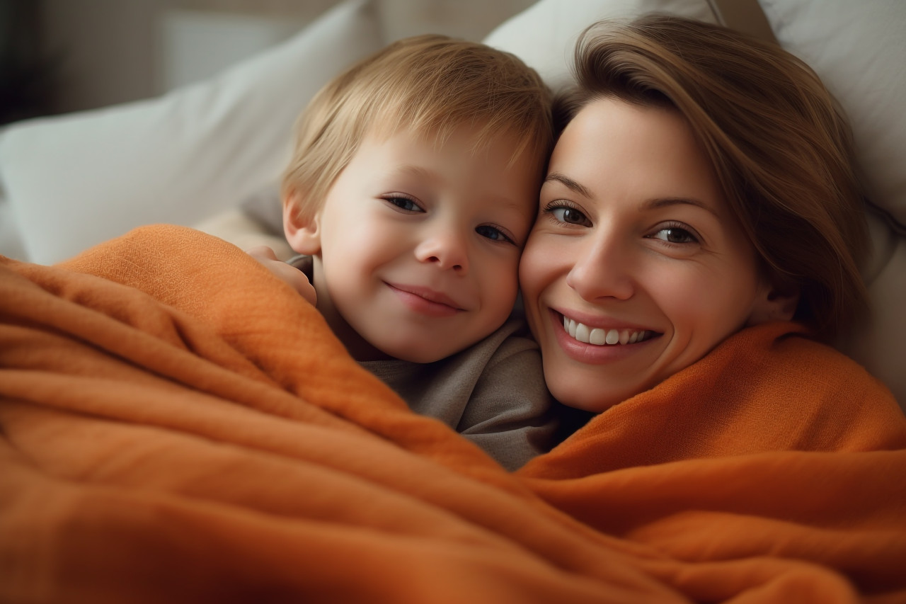 A picture of a mother and her son cuddling under an orange blanket on their bed at home, kids indoor activities