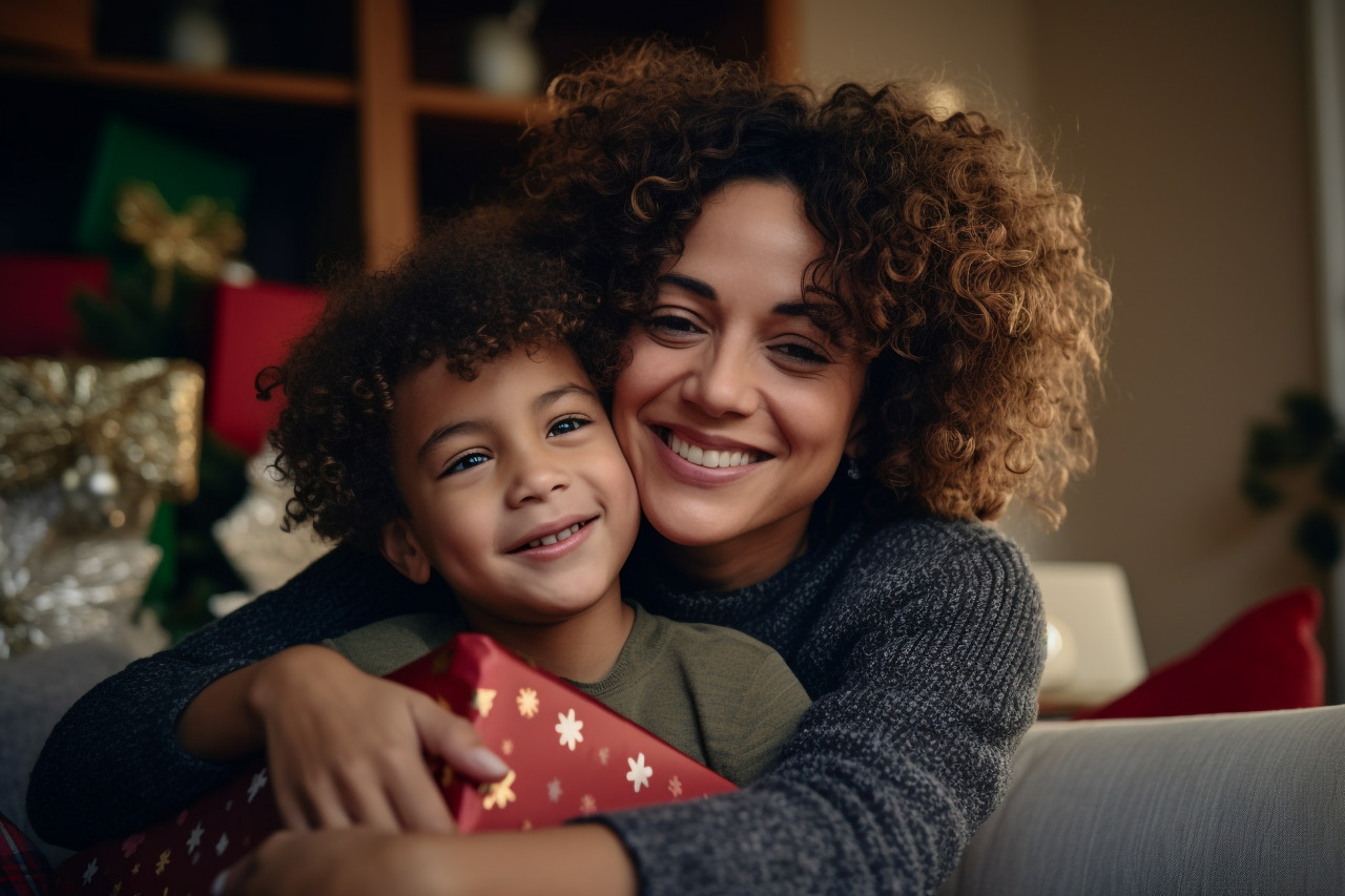 A happy woman of color with a gift is smiling and hugging a child on a couch while celebrating christmas at home, christmas festival people image