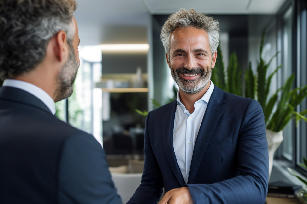 A photo shows a businessman shaking hands with a colleague in an office the businessman is standing and smiling, business and corporate inside office photo
