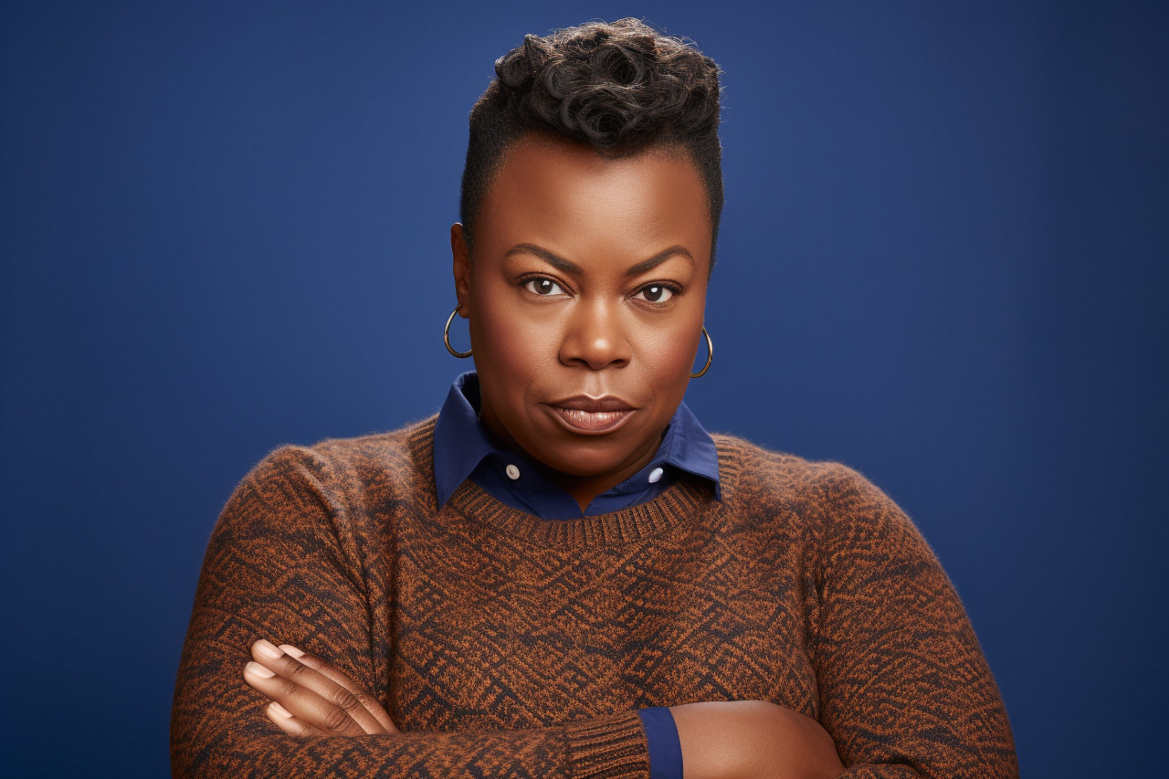 A picture of a serious and plump black woman wearing a blue sweater and standing in front of a plain brown background