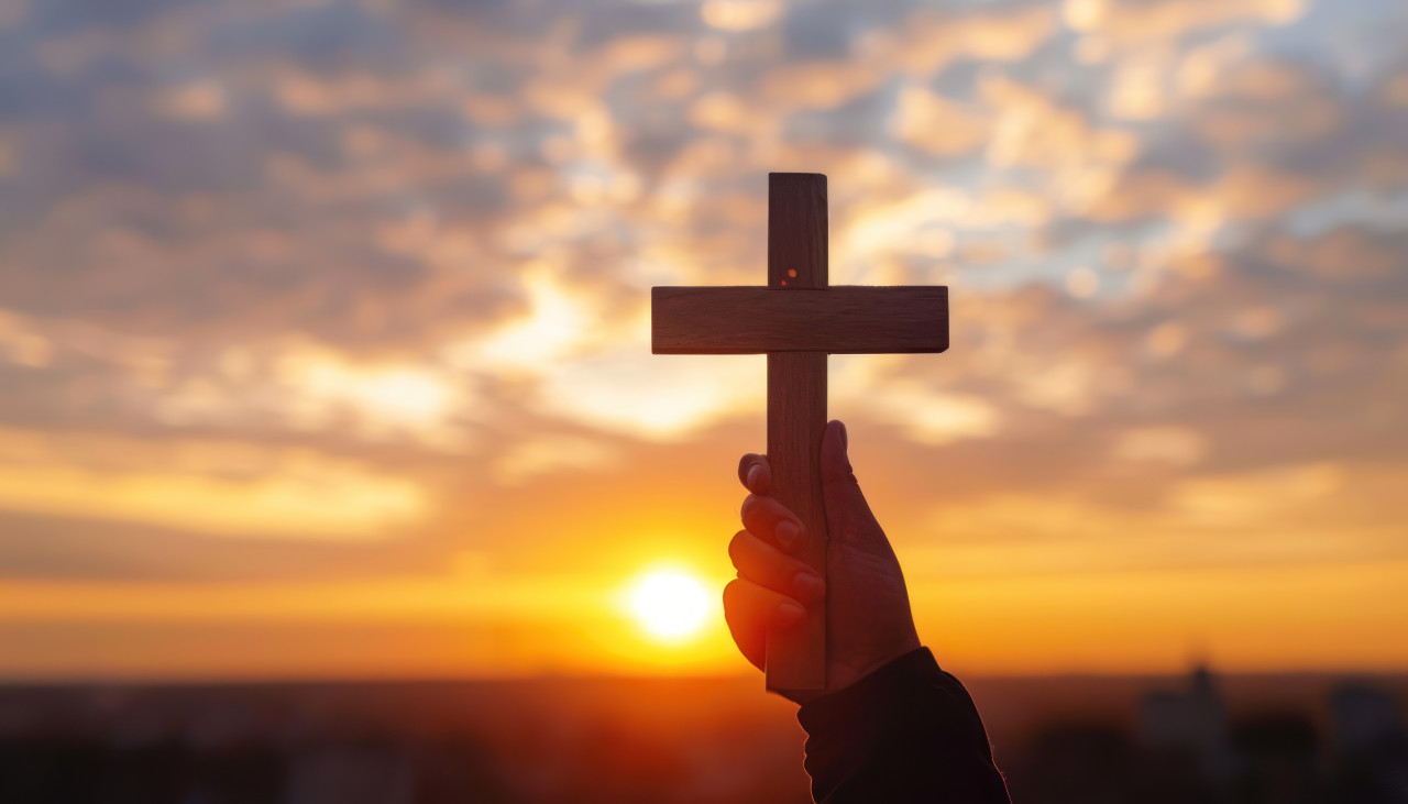 Hand holding cross in silhouette against sunrise backdrop, palm sunday sunset photo