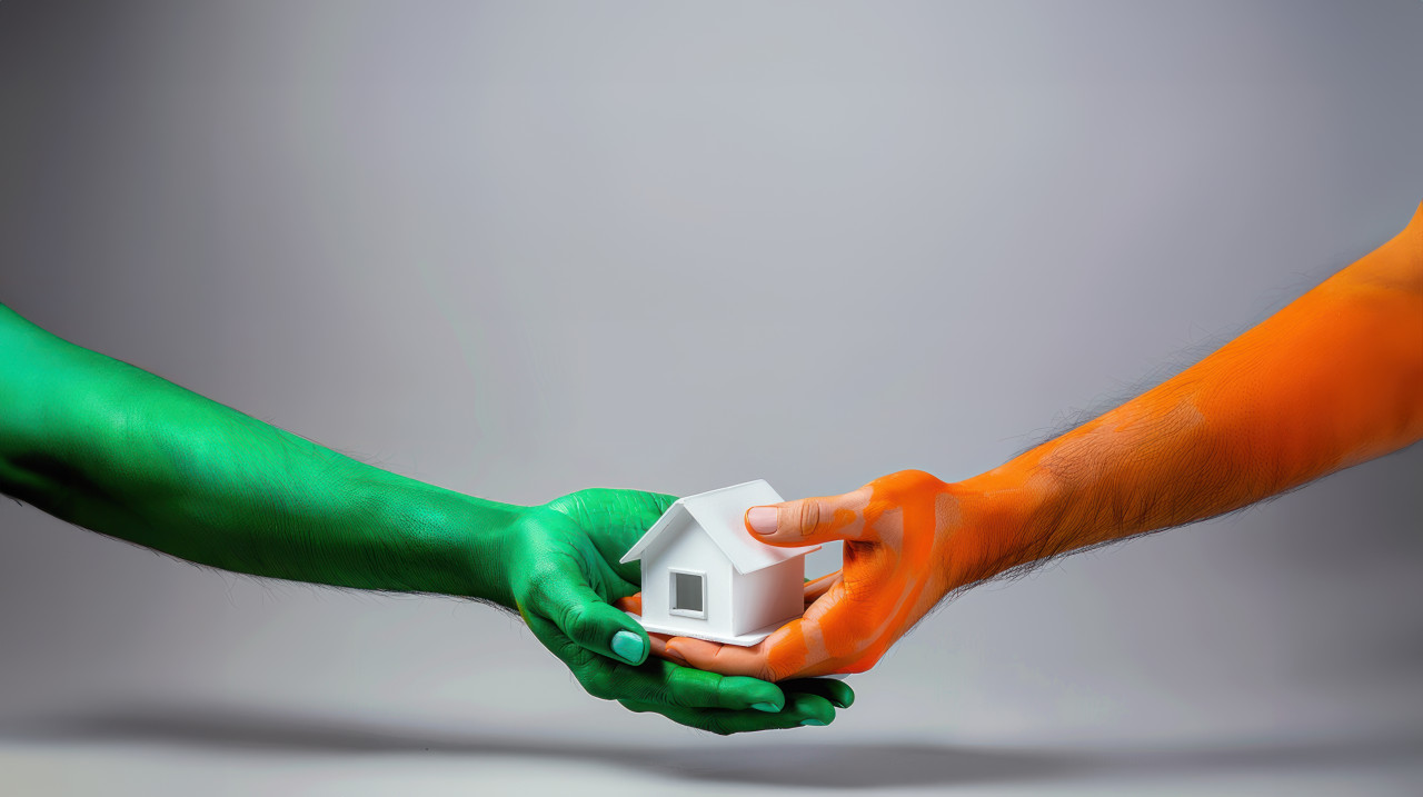 Two hands painted in orange and green colors holding white model house against gray background independence day