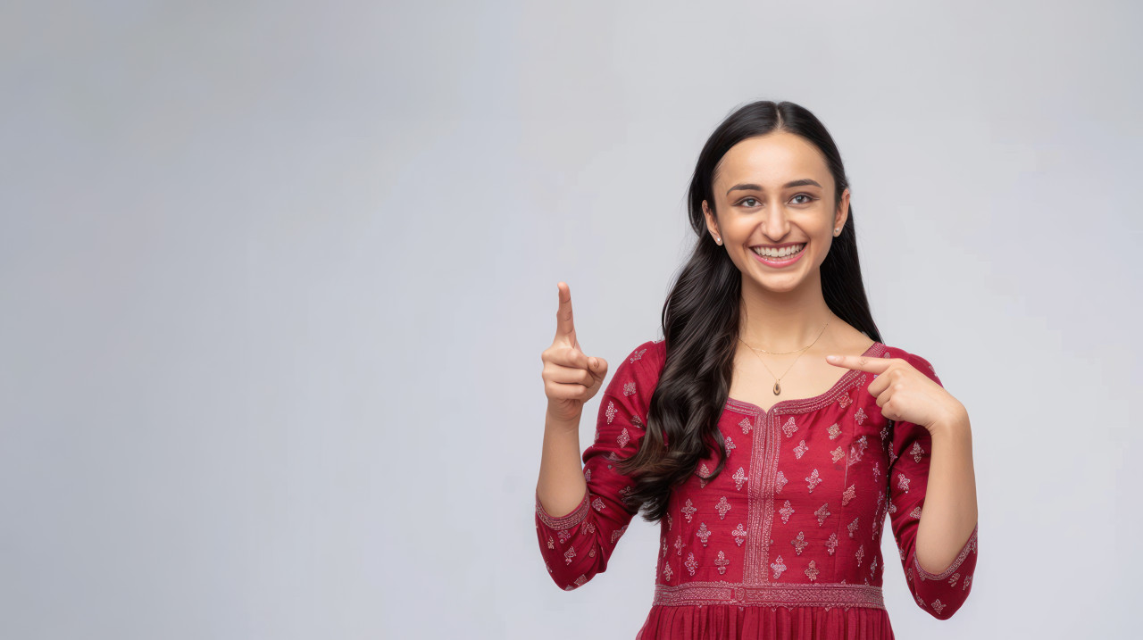 Beautiful indian woman in a red dress smiling and pointing to the right side with her finger standing against a white background, celebrating diwali, festive cheer