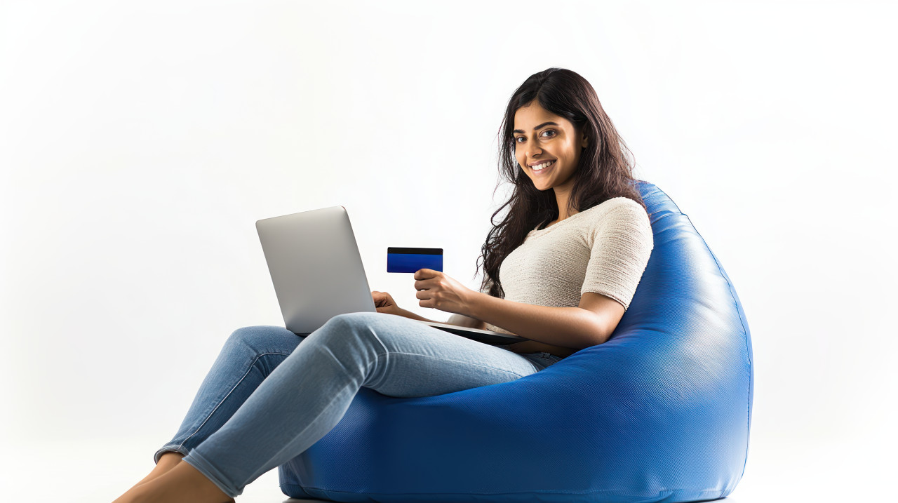 Indian woman sitting on a bean bag holding a credit card and using a laptop atm service theme