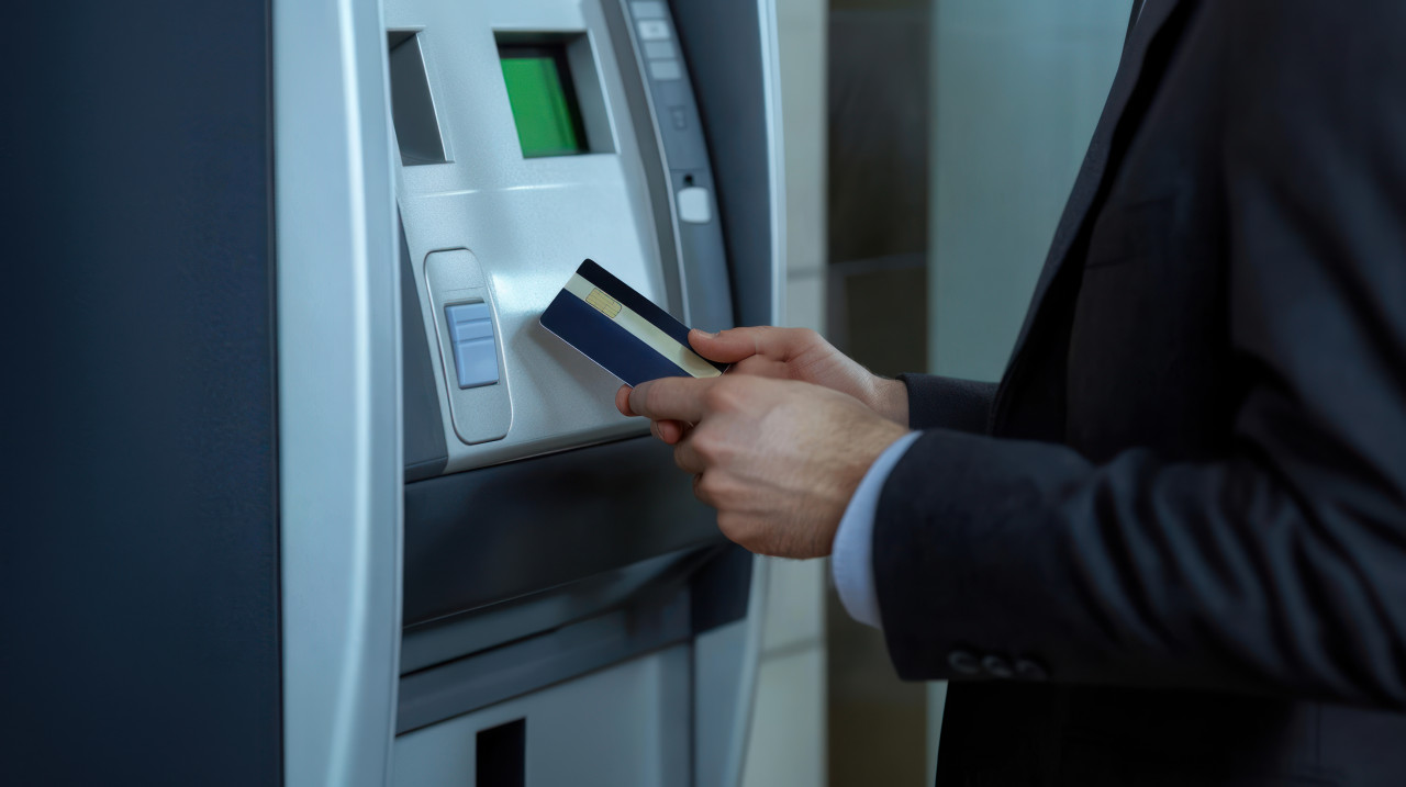 Man using a credit card to make a cash withdrawal at an atm with his smartphone near his hand