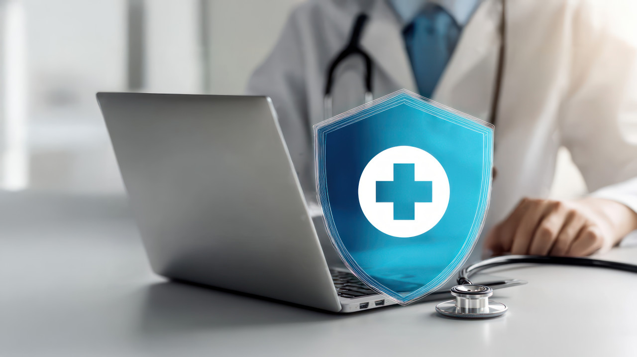 Blue shield with white cross surrounded by medical icons positioned above open laptop, patient security, healthcare data, healthcare security